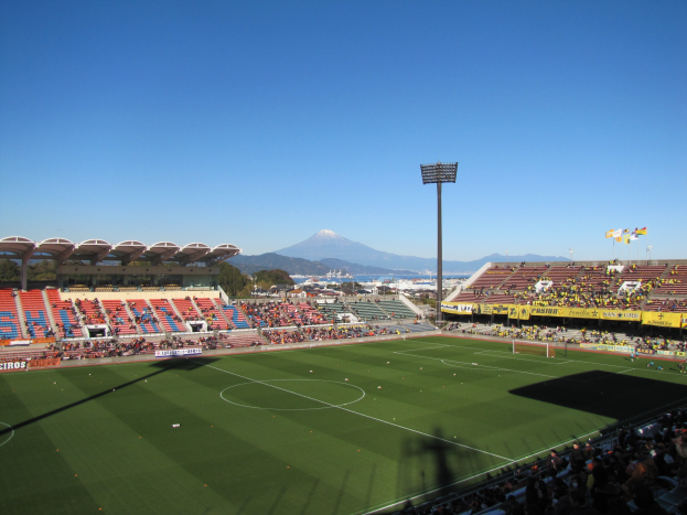 Ein hell erleuchtetes Fußballstadion mit Zuschauern auf den Rängen, umgeben von Bäumen und Hügeln unter einem sichtbaren Himmel, mit Laternenmasten, Bannern und Fahnen.