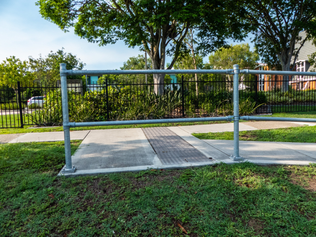 Ein Metallzaun auf einer Rasenfläche, umgeben von Pflanzen, Bäumen und einem Auto auf der Straße, mit Gebäuden und einem klaren blauen Himmel im Hintergrund.