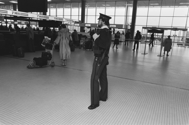 Polizeibeamter in Uniform und Mütze mit einer Waffe in einem Flughafen-Terminal umgeben von Menschen, Gepäckwagen und Gepäck mit Glaswänden und Deckenleuchten im Hintergrund.