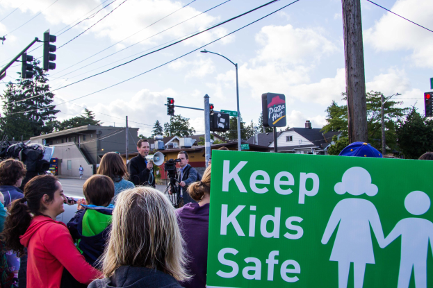 Eine Gruppe von Menschen hält ein "Kinder sicher"-Schild an einer Straßenkreuzung, mit einem Mann mit Megafon im Vordergrund und Gebäuden, Bäumen und Straßen-Elementen im Hintergrund.