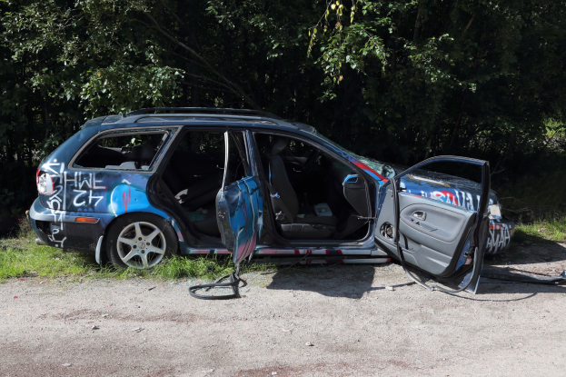 Ein verunfalltes Auto am Straßenrand, mit Gras und Bäumen im Hintergrund.