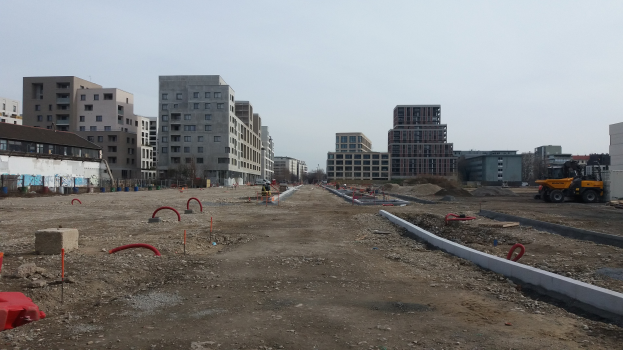 Baustelle mit schwerem Gerät im Vordergrund, Gebäude und Bäume im Hintergrund unter einem klaren blauen Himmel.