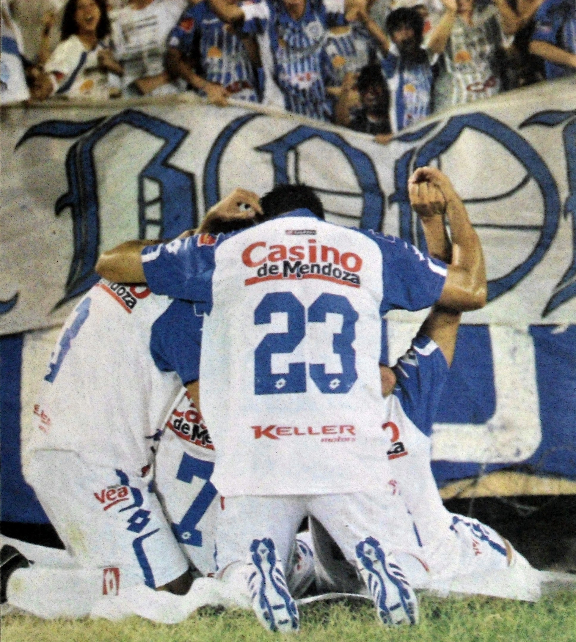 Eine Gruppe von Fußballspielern feiert ein Tor auf dem Boden vor einer jubelnden Menge, mit einem Banner mit Text im Hintergrund.