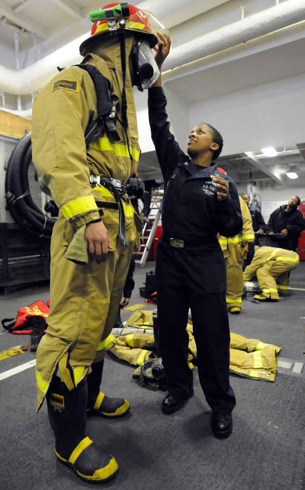 Zwei Feuerwehrleute, eine in schwarzer Uniform und Helm und ein anderer in gelb-schwarzer Ausrüstung, stehen neben einer Leiter und Schläuchen mit anderen im Hintergrund.