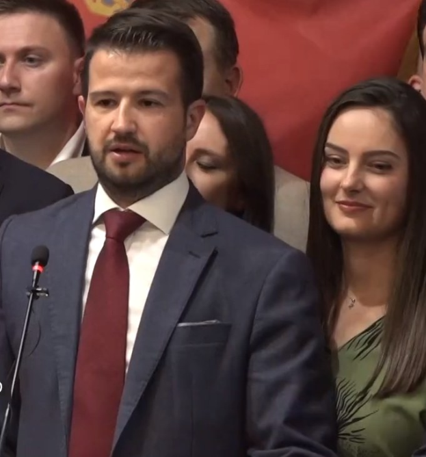 Ein Mann im Anzug spricht an einem Rednerpult mit einem Mikrofon zu einer Menge hinter einer Fahne, wahrscheinlich über ein gouvernementales Verbot von Social Media.