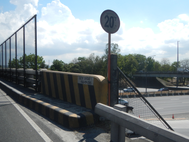 Geschwindigkeitsbegrenzungsschild auf einer Brücke mit Fahrzeugen, einem Zaun, Pfosten, Lampen, Bäumen und einem bewölkten Himmel im Hintergrund.