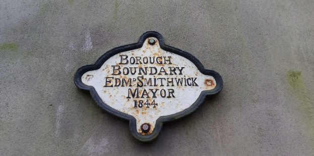 Metallplatte an einer Gebäudeseite mit schwarzem Hintergrund und weißer fettgedruckter Schrift, die "Stadtteilgrenze Edm Smithwick Bürgermeister" liest.