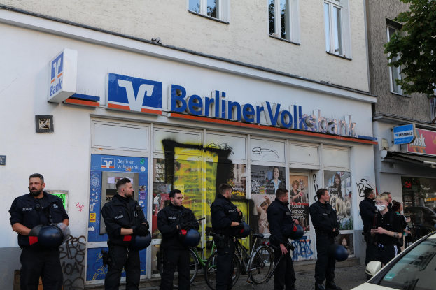 Gruppe von Polizeibeamten vor einer Berliner Volksbank stehend, einige halten Helme in den Händen, mit einem Auto auf der rechten Seite und einem Baum auf der linken Seite, Gebäude und Texttafeln im Hintergrund.