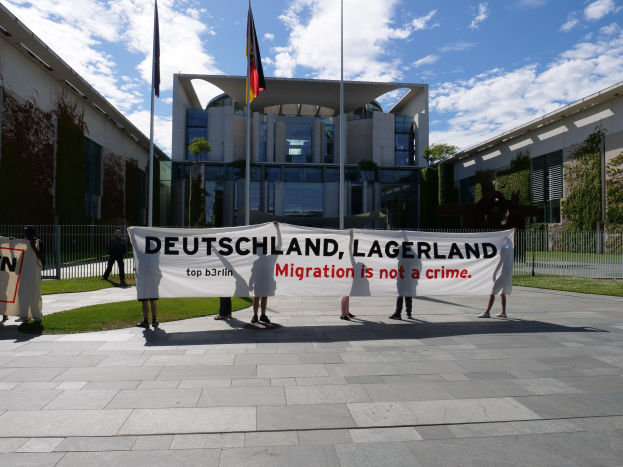 Gruppe von Menschen mit einer Tafel "Deutschland, Lagerland Migration ist kein Verbrechen" vor einem Gebäude mit Fenstern, umgeben von Gras, einem Metallzaun, Pflanzen, Bäumen und Fahnen auf Ständen unter einem bewölktem Himmel.