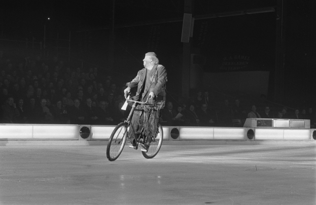 Schwarze und weiße Fotografie eines Mannes, der auf einem Eisstadion Fahrrad fährt, mit einer Gruppe von Sitzenden im Hintergrund und einer Wand dahinter.