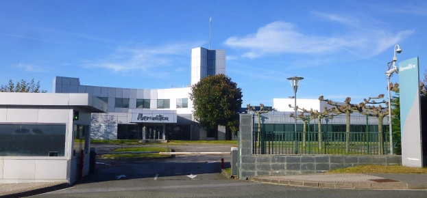 Ein großes, mehrfenstriges Gebäude mit einem Tor davor, umgeben von Straßenmasten, Laternen, Gittern, Bäumen und Gras, unter einem bewölkten Himmel, das als Hauptquartier von Iberostica in den Niederlanden dient.