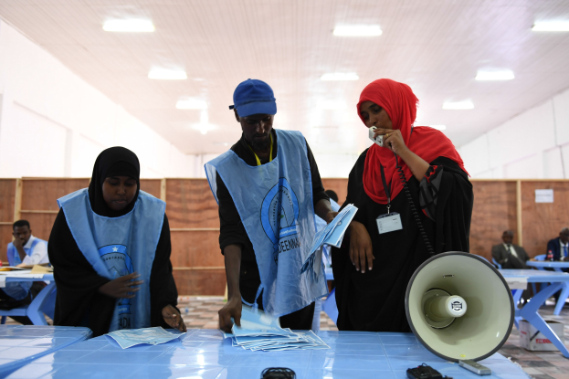 Menschen versammeln sich um einen Tisch mit einem Megafon und Papieren an einer Wahllokal, mit anderen im Hintergrund auf Stühlen sitzend und Stehenden unter Deckenbeleuchtung.
