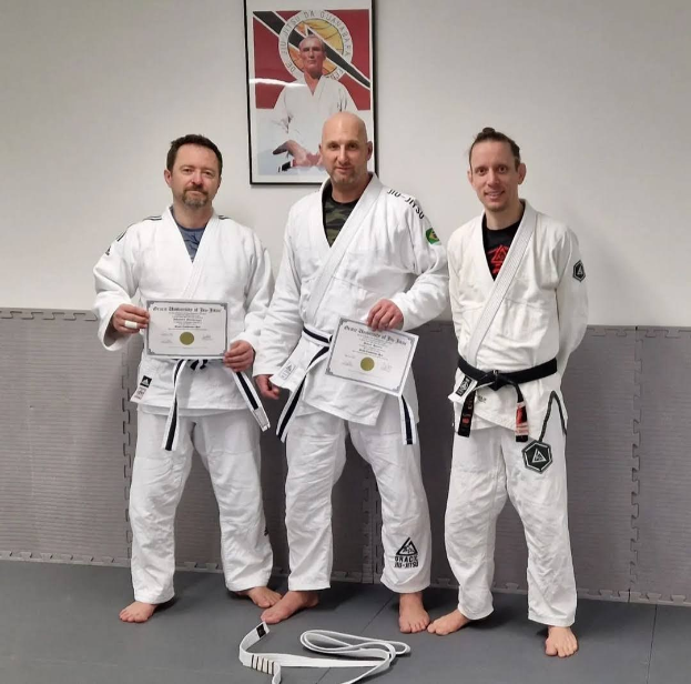 Drei Männer in weißen Judo-Unterhemden mit Urkunden, die in einem Fitnessstudio mit einem gerahmten Preis im Hintergrund stehen.