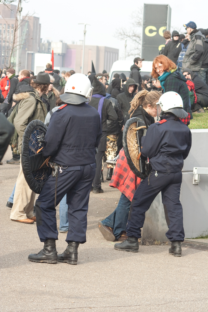 Eine Gruppe von Menschen auf einer Straße mit zwei Personen vorne, die wie Polizei aussehen, Gebäuden im Hintergrund und Boden unten.