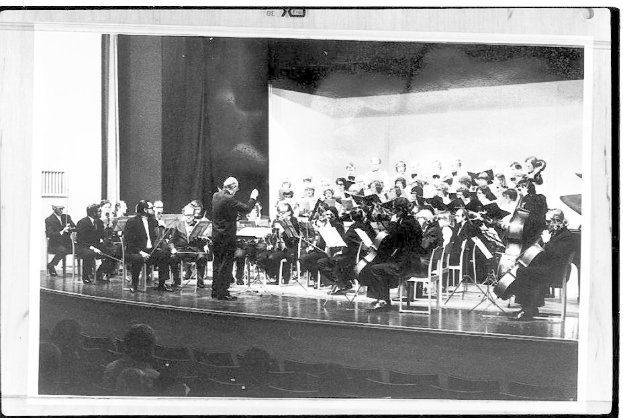 Ein Schwarz-Weiß-Foto eines Dirigenten in Anzug und Krawatte, der ein Orchester mit Geigen, Celli und Flöten leitet, während das Publikum aufmerksam lauscht.