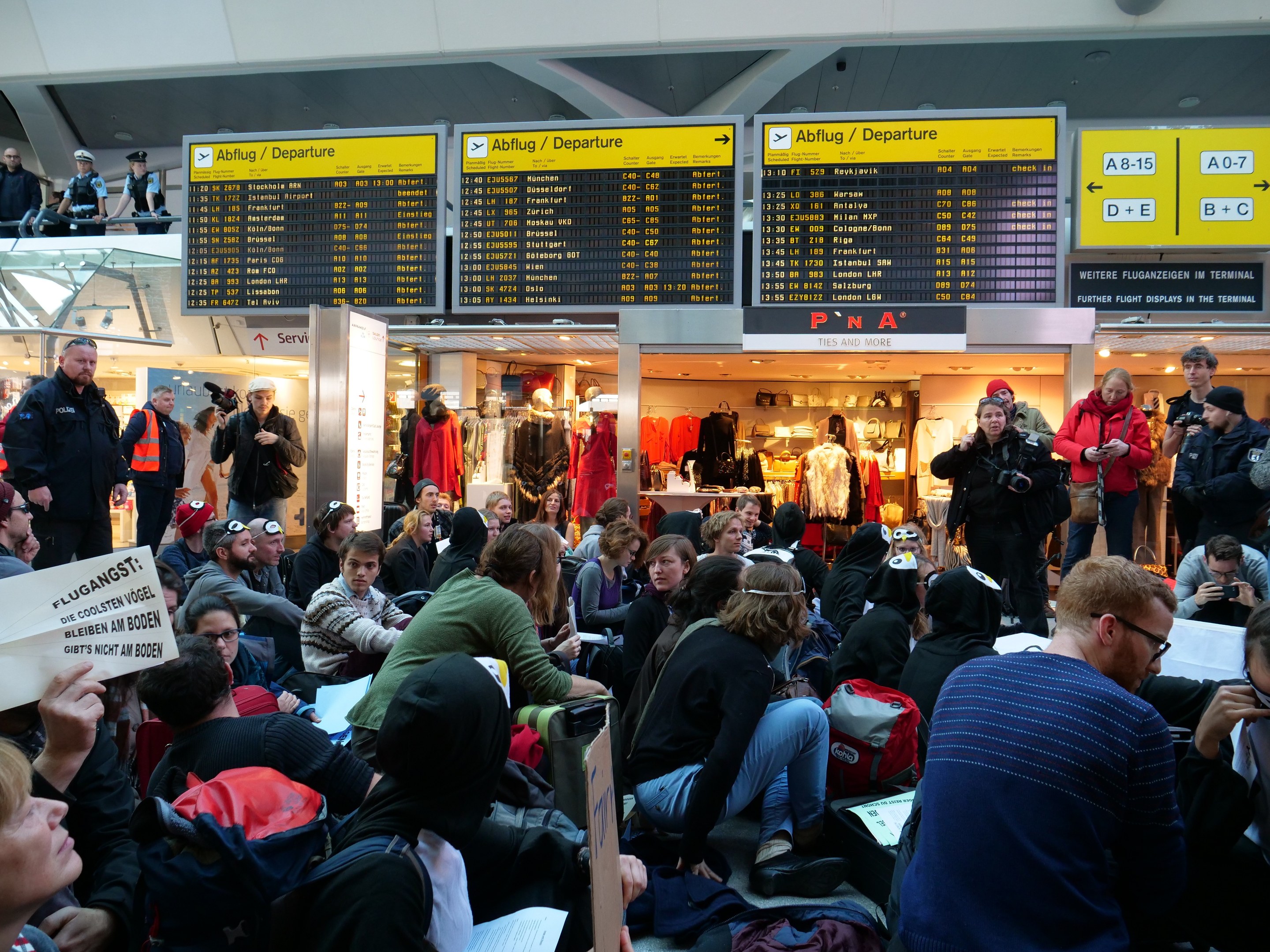Eine große Gruppe von Menschen in einem Flughafen, einige mit Taschen und Papieren sitzend, andere stehend, mit Texttafeln, Schaufensterpuppen in Kleidern und Deckenlampen im Hintergrund, was auf eine Demonstration hinweist.