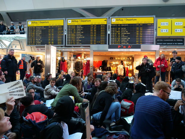 Eine große Gruppe von Menschen in einem Flughafen, einige mit Taschen und Papieren sitzend, andere stehend, mit Texttafeln, Schaufensterpuppen in Kleidern und Deckenlampen im Hintergrund, was auf eine Demonstration hinweist.
