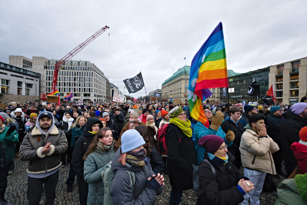 Eine gro├če Gruppe von Menschen bei einer LGBTQ+-Rechte-Demonstration in Berlin, die Fahnen und Plakate schwenken, mit Geb├Ąuden, einem Kran und Wolken im Hintergrund.