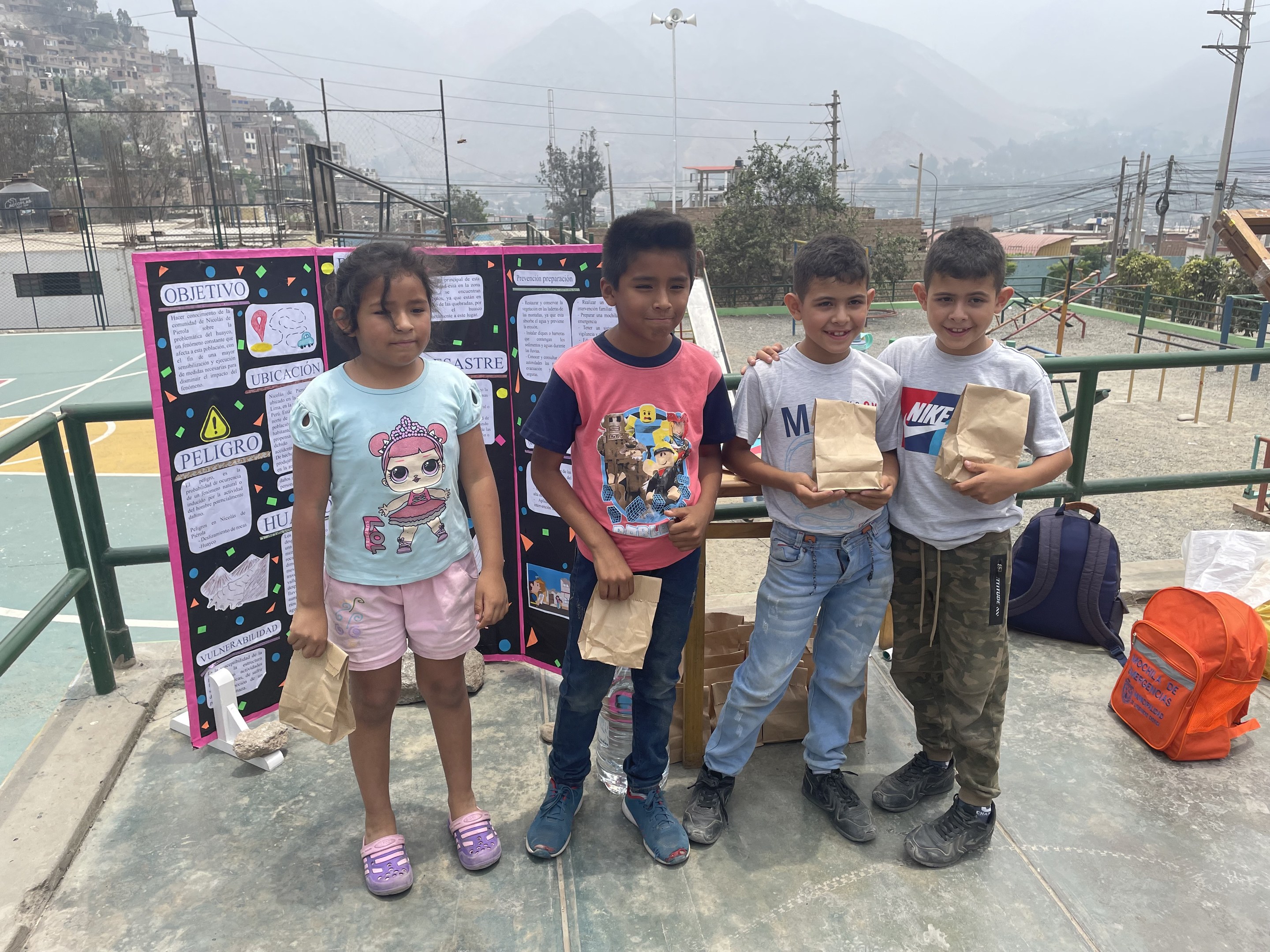 Gruppe von Kindern auf einem Basketballfeld mit Papiertüten, mit Vorräten auf dem Boden und einer Tafel mit Text links daneben, vor einem Hintergrund aus Zaun, Pfosten, Straßenlaternen, Bäumen, Gebäuden, Hügeln und Himmel.