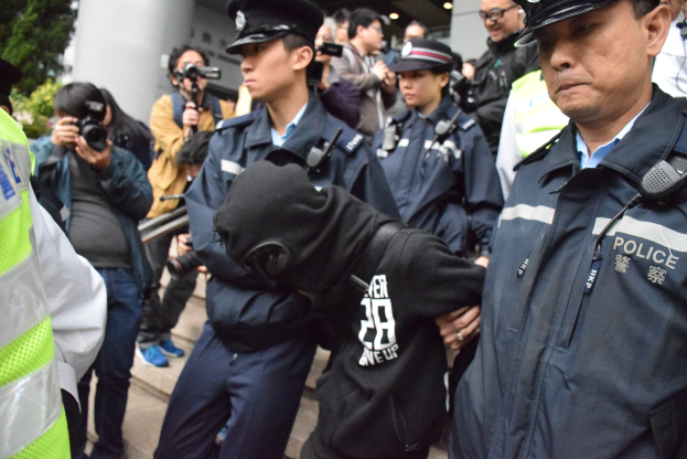 Eine Gruppe von Polizeibeamten steht vor einer Menge vor einem Gebäude mit Säulen, mit Bäumen auf der linken Seite.
