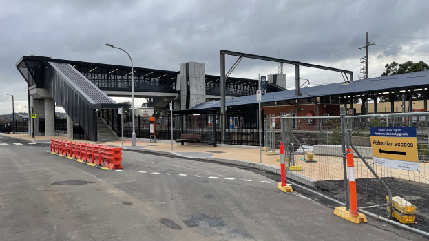 Ein Bahnhof mit einer Brücke im Hintergrund, Straßenlaternen, Strommasten, Stromkabel, Bäumen, Gebäuden, Kraftfahrzeugen auf der Straße, Absperrpoller, Schilder, Bänken und einem bewölkten Himmel.