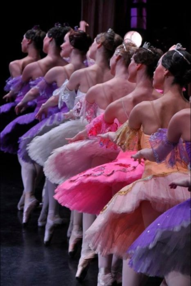 Eine Gruppe von Balletttänzerinnen in rosafarbenen Tutus sitzt auf einer Bühne, beleuchtet von einem Scheinwerfer im Hintergrund, der sich im Einklang bewegt.