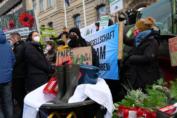 Eine Gruppe von Menschen in Masken mit Protestschildern vor einem Lkw mit einem Tisch voller Gegenstände, Topfpflanzen und Gebäuden im Hintergrund, die gegen ein Pestizidverbot protestieren.