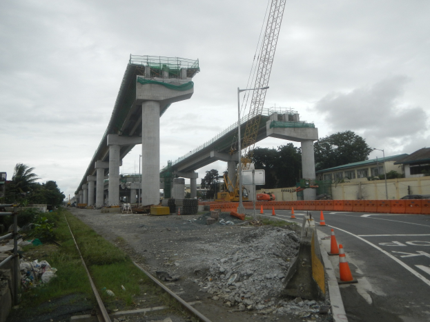 Baustelle mit einer Brücke im Hintergrund, Straße mit Verkehrskegeln markiert, verstreute Steine und Gras, Eisenbahnschiene links, Bäume und Gebäude auf beiden Seiten der Straße und ein bewölkter Himmel oben.