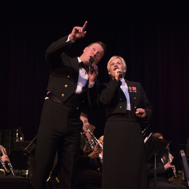 Ein Mann und eine Frau in Smokings stehen auf einer Bühne und halten Mikrofone, hinter ihnen sitzt ein Orchester.