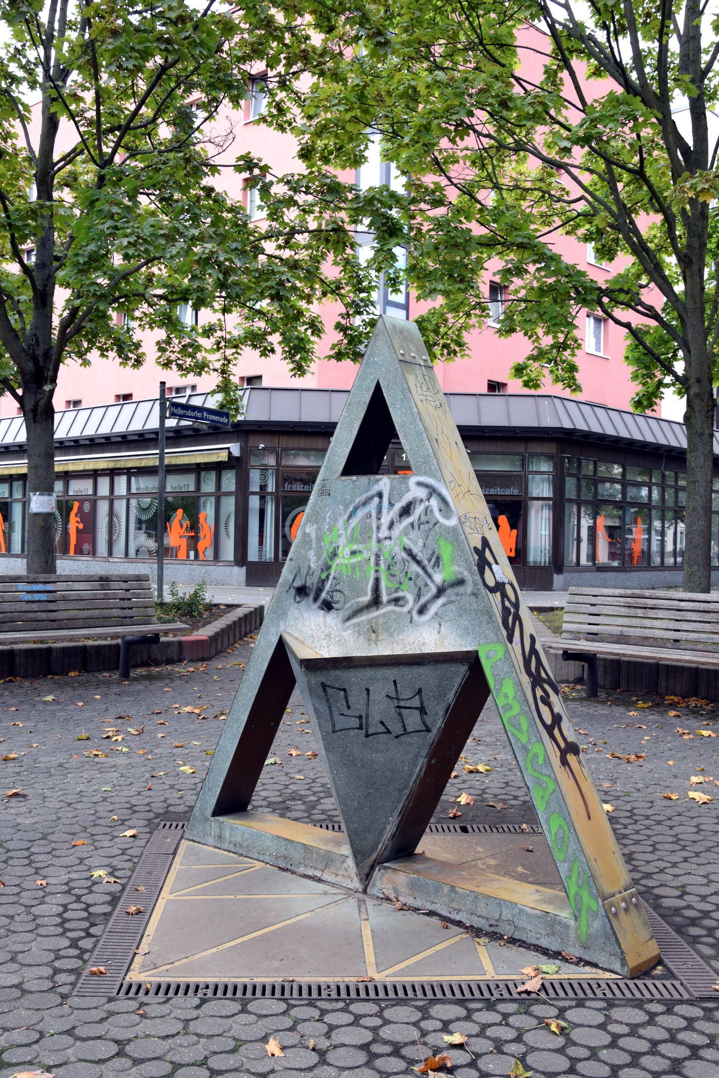 Metallskulptur mit Graffiti in einem Park, umgeben von Bäumen, Bänken, einem Pfahl und einem Gebäude im Hintergrund.