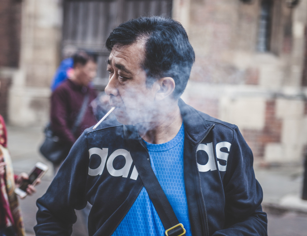 Ein Mann in einem blauen T-Shirt und schwarzer Jacke raucht eine Zigarette auf der Straße, während andere im Hintergrund gehen, einer hält ein Mobiltelefon, mit einem Gebäude im Hintergrund sichtbar.