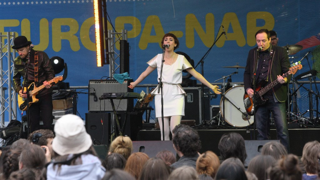 Vier Musiker spielen auf der Bühne: Eine Frau in einem weißen Kleid singt in der Mitte, während zwei Männer links und rechts von ihr Gitarre spielen, mit einer sichtbaren Menge unten und Lautsprechern an der Wand im Hintergrund.