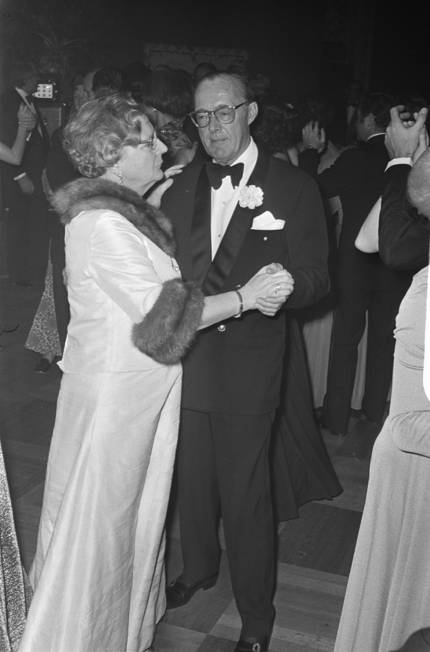 Ein Mann in einem schwarzen Blazer, weißem Hemd und schwarzen Hosen tanzt mit einer Frau in einem eleganten Kleid auf einer Party, mit anderen Menschen und Kameras im dunklen Hintergrund.