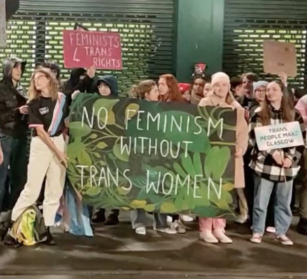 Eine Gruppe Von Menschen Hält Eine Banner Mit Der Aufschrift "Kein Feminismus Ohne Trans-Frauen" Und Verschiedene Plakate In Einer Öffentlichen Räumlichkeit.