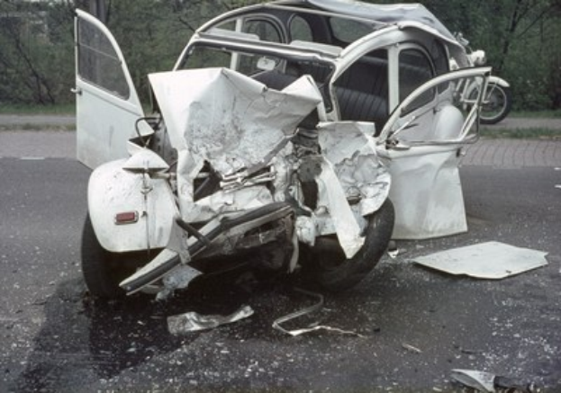 Schwer beschädigt Frontpart eines Autos am Straßenrand mit Bäumen und Gebäude im Hintergrund, weiteres Fahrzeug in der Ferne sichtbar.