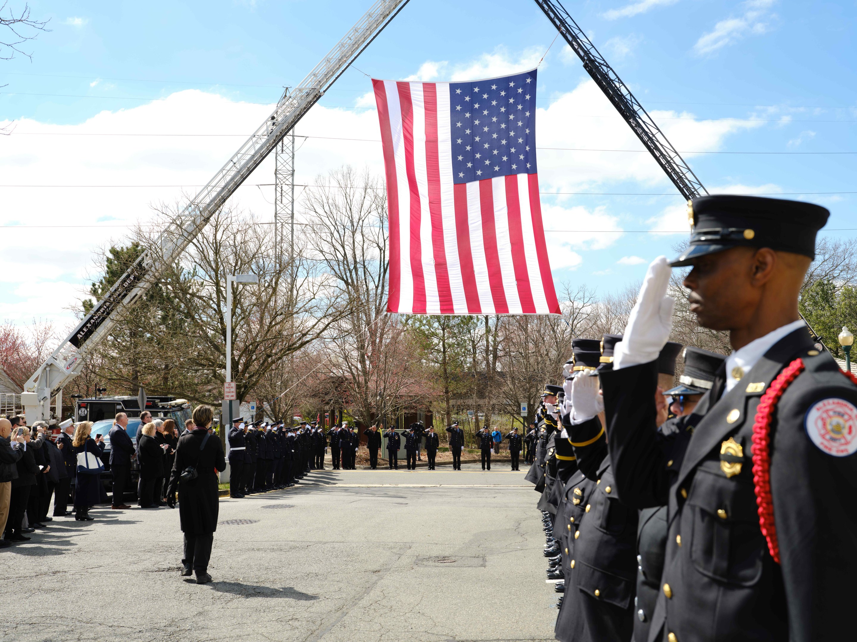 Gruppe uniformierter Personen vor einem Feuerwehrauto stehend, die grüüend und eine amerikanische Flagge haltend, mit Bäumen, Gebäuden und Laternenmästen im Hintergrund.