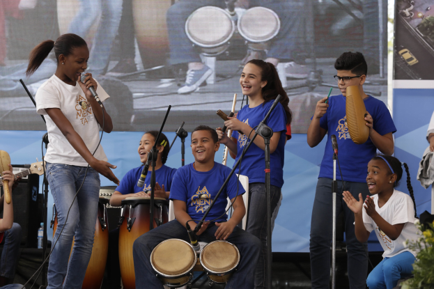 Gruppe von Kindern, die auf einer Bühne Musikinstrumente spielen, mit einer Frau in der Mitte, die ein Mikrofon hält, umgeben von Mikrofonstativen, einem Banner im Hintergrund und Pflanzen auf der rechten Seite.