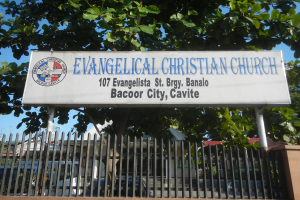 Eine evangelische Kirche in der Stadt Bacoor, Cavite, mit einem Schild, einem Metallzaun, einer Wand, Bäumen, Fahrzeugen, Gebäuden, Pfählen und einem bewölkten Himmel.