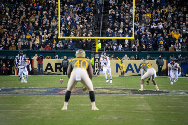 Fußballspiel in einem Stadion mit Spielern in Sportkleidung und Helmen im Vordergrund und Zuschauern im Hintergrund sowie sichtbaren Stadionwerbung und Pfosten.
