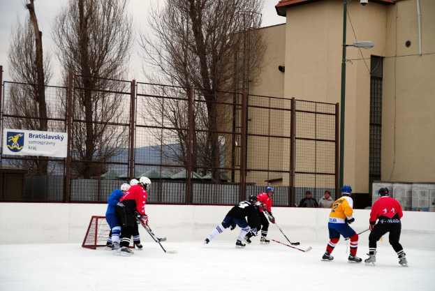 Menschen, die Eishockey auf einem Eisplatz mit Gebäuden, Bäumen, einer Straßenlaterne, einem Namensschild und Zäunen im Hintergrund unter einem klaren Himmel spielen.