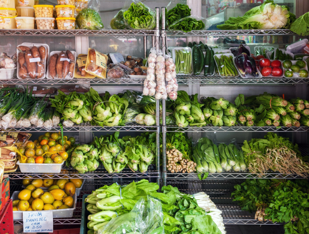 Ein Gang in einem Supermarkt mit frischem Gemüse und Obst in Plastikverpackungen, Kisten und einer Texttafel sowie einem Glasfenster im Hintergrund.
