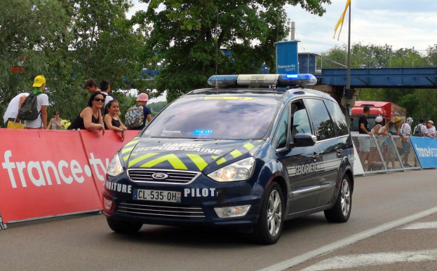 Polizeiauto fährt an einer Menschenmenge vorbei, mit einer Fahne, Bäumen, einer Brücke, einer Flagge und einem bewölkten Himmel im Hintergrund.