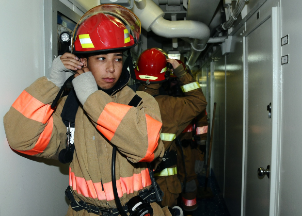 Feuerwehrleute in Uniform, die in einem Raum mit Rohren und Gegenständen im Hintergrund stehen, wahrscheinlich während einer Übung.