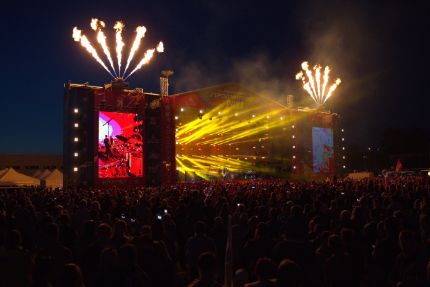 Eine große Menschenmenge bei einem Musikfestival schaut auf eine hell erleuchtete Bühne mit Bannern, umgeben von Zelten und Feuerwerk am Nachthimmel.