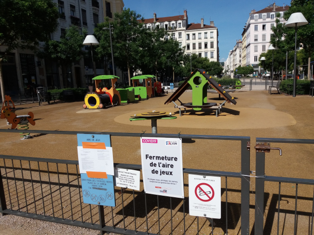 Ein eingezäunter städtischer Spielplatz mit mehreren Schildern am Zaun, umgeben von Bäumen, Laternenmasten und Gebäuden unter einem sichtbaren Himmel.