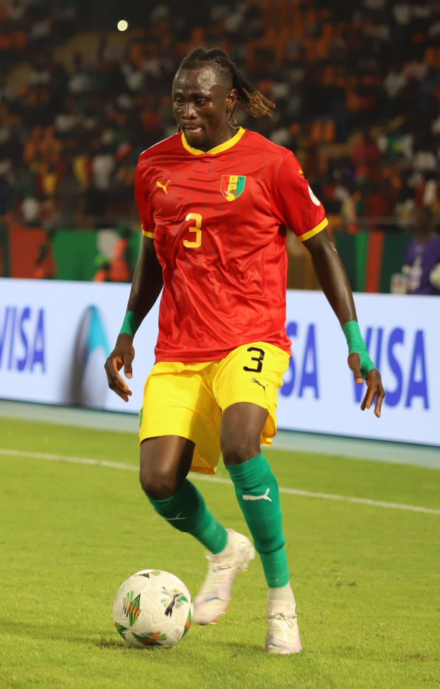 Ein Mann in rot-gelber Uniform spielt Füßball auf einem Feld, umgeben von Zuschauern auf den Tribünen und Werbetafeln im Hintergrund.