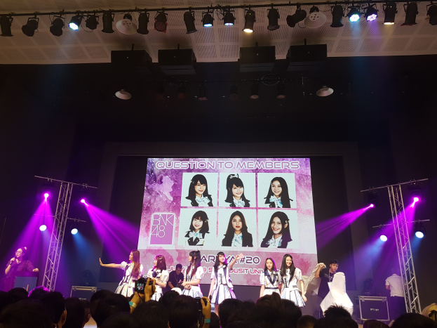 Eine Gruppe von Menschen performs auf einer hell beleuchteten Bühne mit Trägern, während ein Sänger ein Mikrofon hält und ein anderer ein Instrument spielt, während die Menge darunter das Konzert aufzeichnet; ein großer Bildschirm im Hintergrund zeigt Bilder der K-Pop-Gruppe BK48.