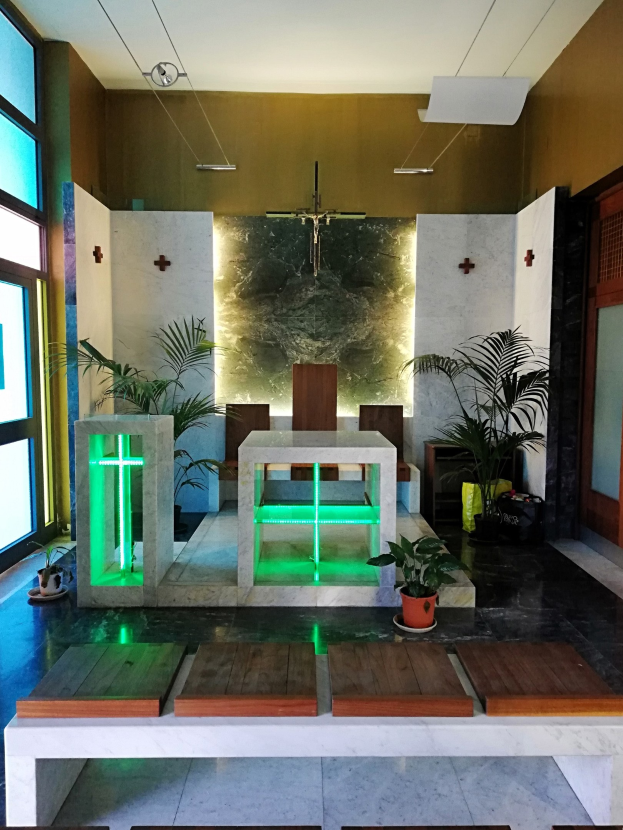Innenseite einer Kirche mit Holzliegen, Zimmerpflanzen, einem Kreuz an der Wand, einer Tür auf der rechten Seite und Glasfenstern auf der linken Seite, beleuchtet von grünem Licht.