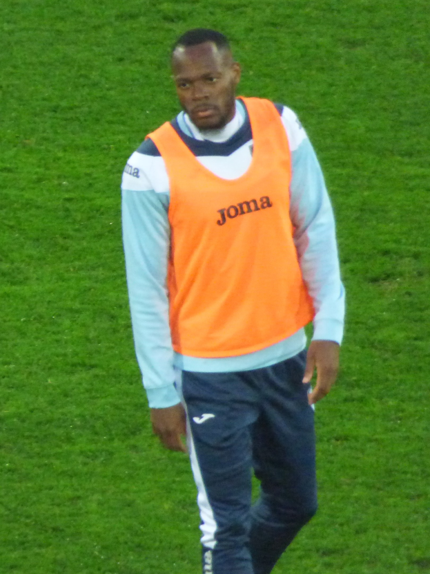 Ein Fußballer in einem orangen Hemd und blauen Hosen, der entschlossen über ein grünes Feld läuft.