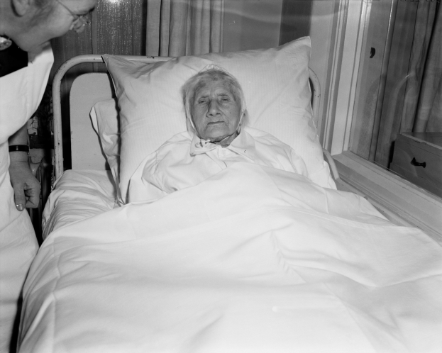 Ein älteres Weib liegt in einem Krankenhausbett mit einer Schwester daneben, das Bett mit einem weißen Laken und Kissen bedeckt und ein Fenster mit Vorhängen im Hintergrund.